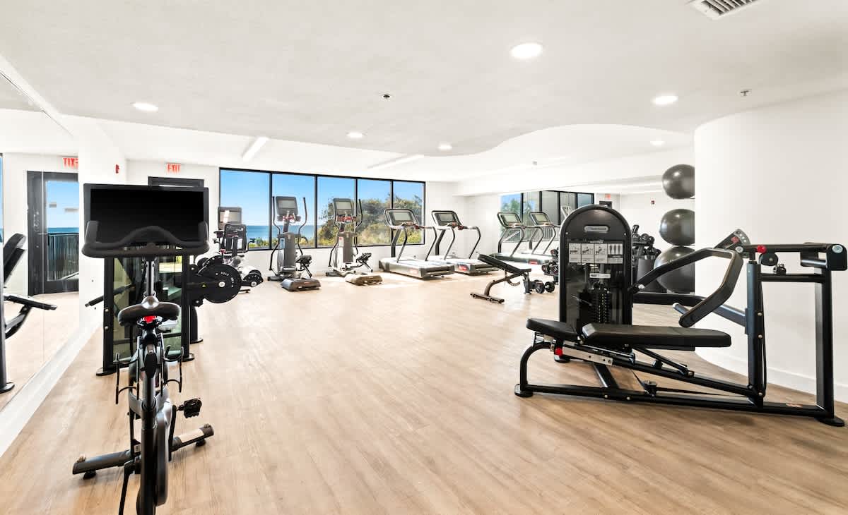 A well-equipped fitness center features a variety of cardio machines like treadmills and stationary bikes, along with weight training equipment. Large windows offer views of the outdoors.