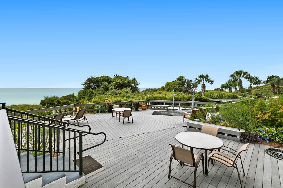 An expansive outdoor deck with gray composite decking features multiple round dining tables with chairs and lounge seating, surrounded by lush green foliage and offering panoramic ocean views.
