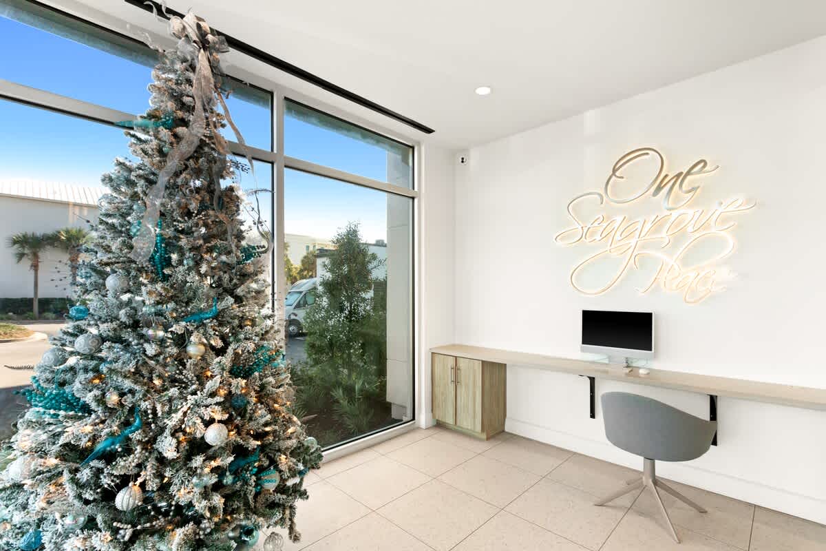 A festive corner of the lobby features a large Christmas tree decorated with blue and silver ornaments next to a window. A minimalist desk with a computer and a gray office chair is also present, beneath a neon sign.