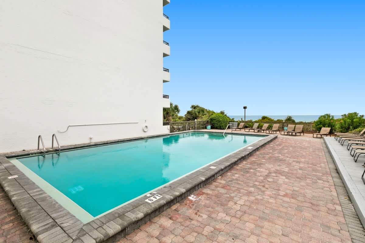 Rectangular outdoor swimming pool with clear blue water, surrounded by a brick paver deck and a white multi-story building, with the ocean visible in the distance.