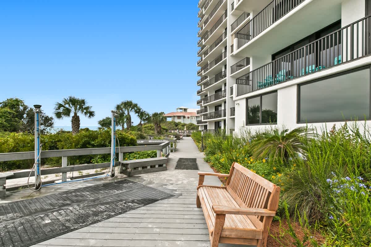 A long wooden walkway with a railing runs alongside a tall white building with balconies, featuring lush green landscaping, outdoor showers, and a wooden bench, leading towards the ocean in the distance.