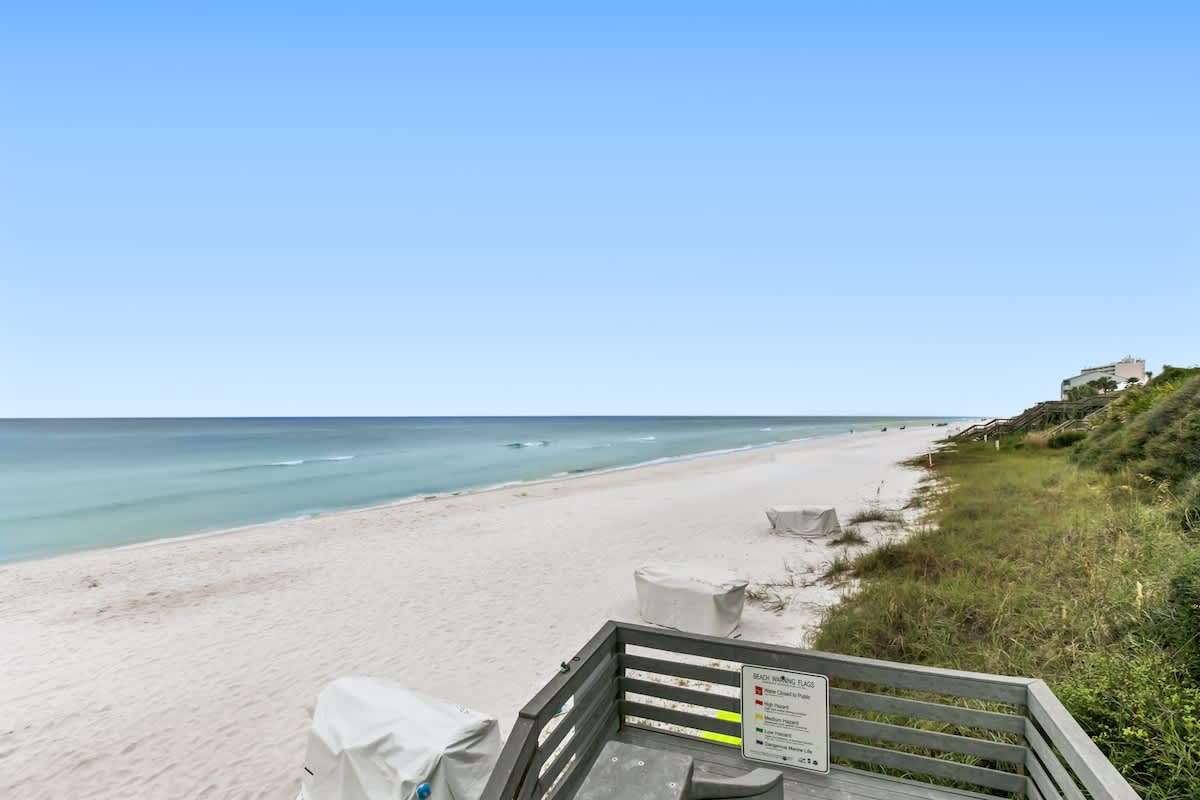 A pristine white sand beach stretches along the turquoise ocean under a clear blue sky, with gentle waves lapping the shore and a wooden boardwalk leading down to the sand.