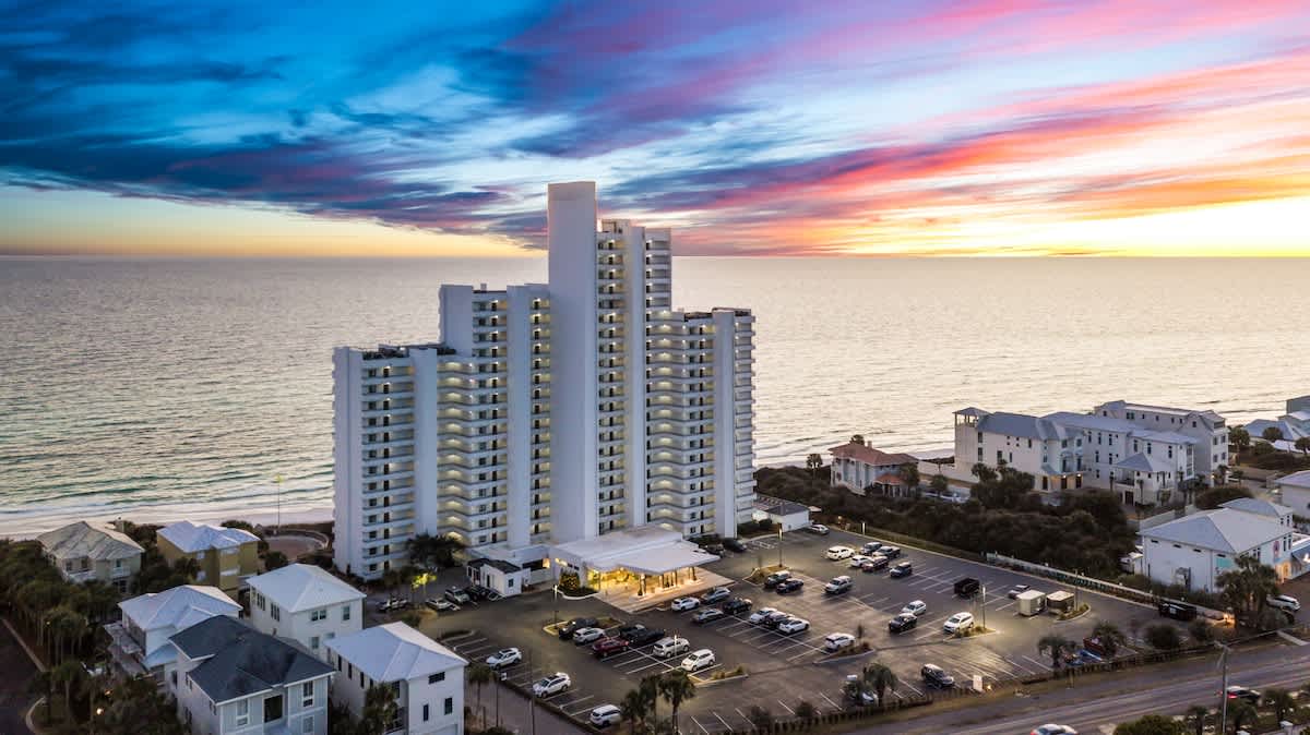 Aerial view of a tall, white, multi-story condominium building situated directly on the coastline, with a large parking lot in the foreground, all under a vibrant sunset sky over the ocean.