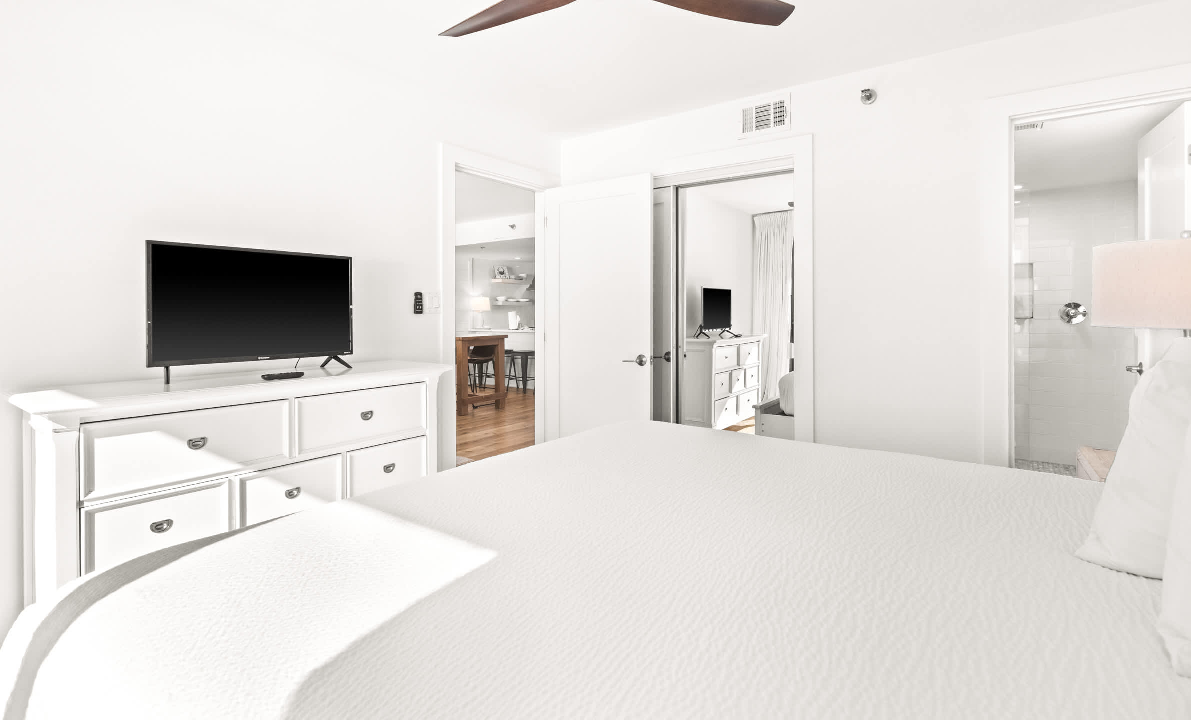 View from the bedroom showing a white dresser with a TV, with an open doorway leading to the kitchen and living area, and another door to the bathroom.