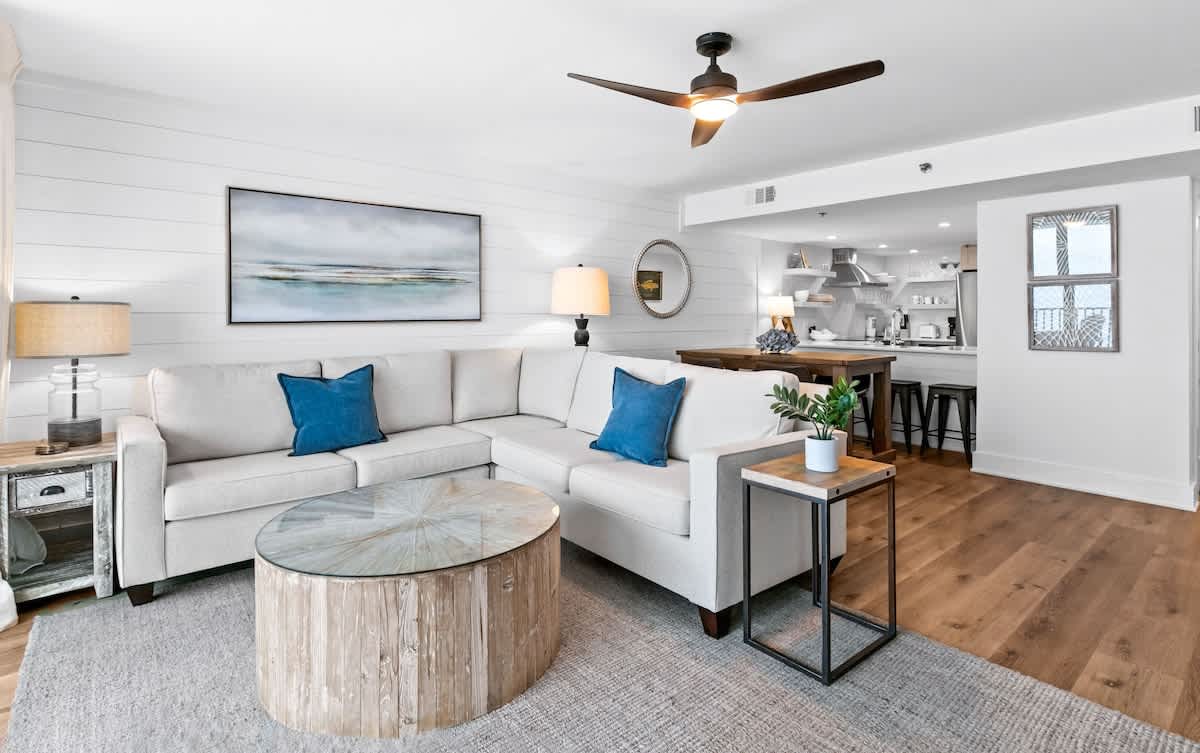 The open-concept living area features a large L-shaped sofa with blue accent pillows, a round wooden coffee table, and a shiplap accent wall, flowing into a modern kitchen with a breakfast bar.