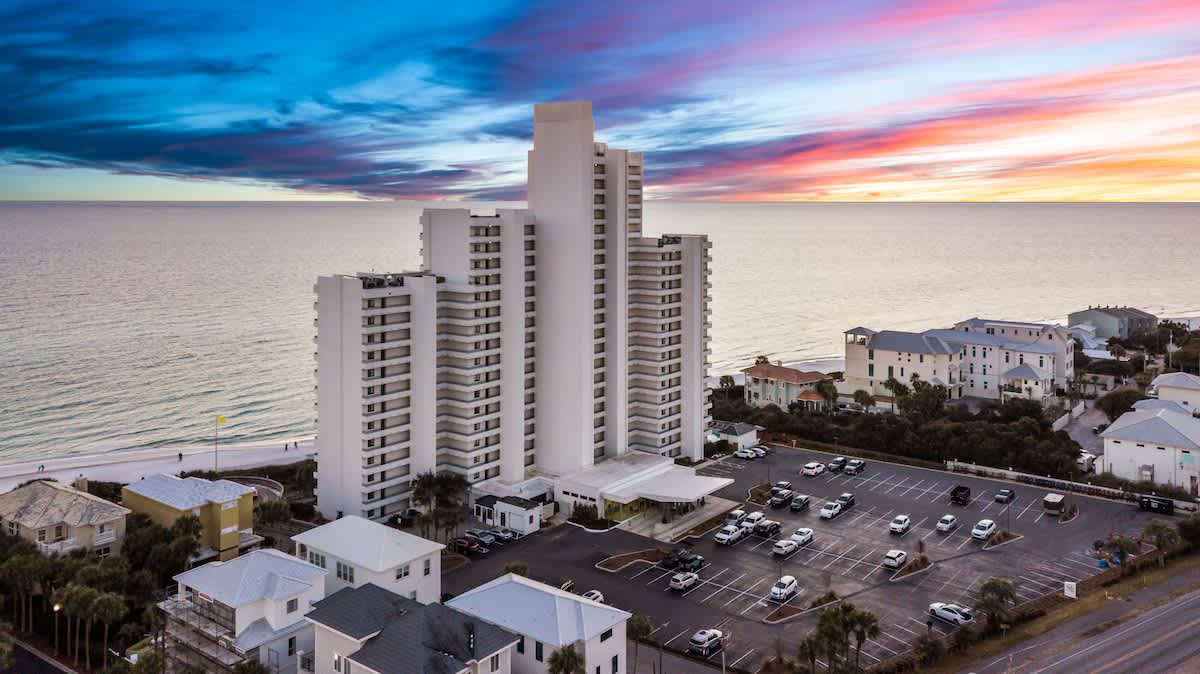 Aerial view of a prominent white high-rise condominium building overlooking the ocean and a sandy beach, surrounded by smaller residential structures and a spacious parking lot, under a dramatic sunset sky.