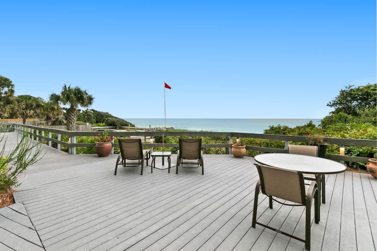 A spacious gray composite deck provides stunning ocean views, featuring a round dining table with chairs and two lounge chairs, all set against a backdrop of green foliage and a distant red flag.