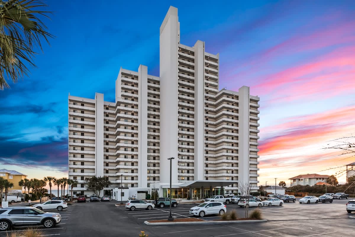 A tall, modern white condominium building with numerous balconies stands against a vibrant sunset sky with hues of blue, pink, and orange. A large parking lot filled with cars is visible in the foreground.