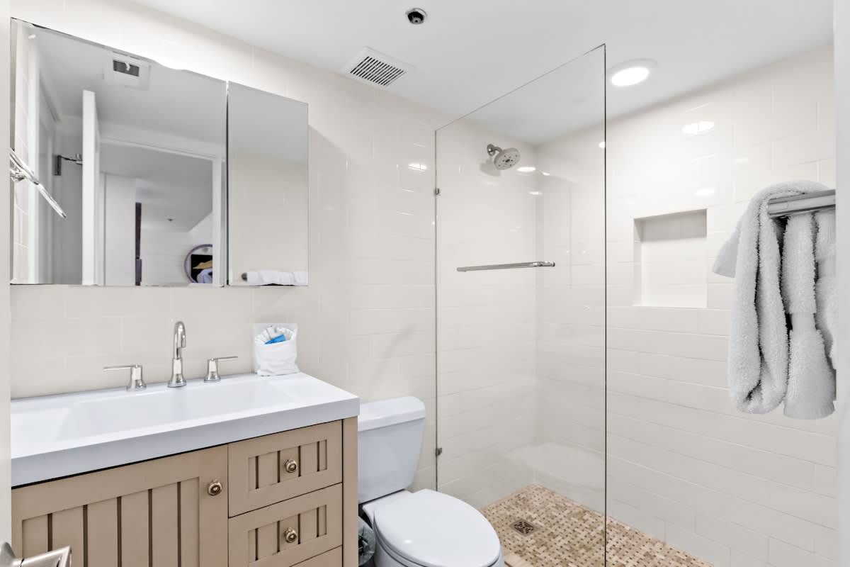 Clean bathroom with a light wood vanity, white sink, and a glass-enclosed walk-in shower with white subway tiles and a woven mat, viewed from a slightly different angle.