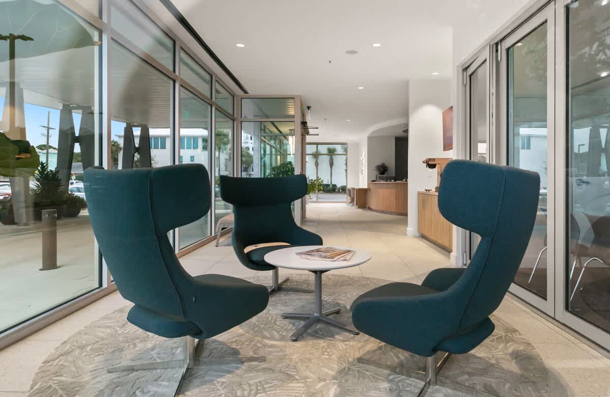 A cozy seating arrangement in the lobby features three modern, high-backed teal armchairs surrounding a small white coffee table on a light-colored patterned rug. Large windows provide natural light.