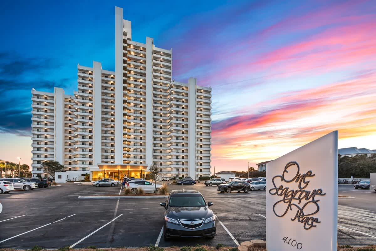 A multi-story white condominium building with numerous balconies is illuminated by the colorful sunset sky. A spacious parking lot with several cars and a large "One Seagrove Place" sign are visible in the foreground.