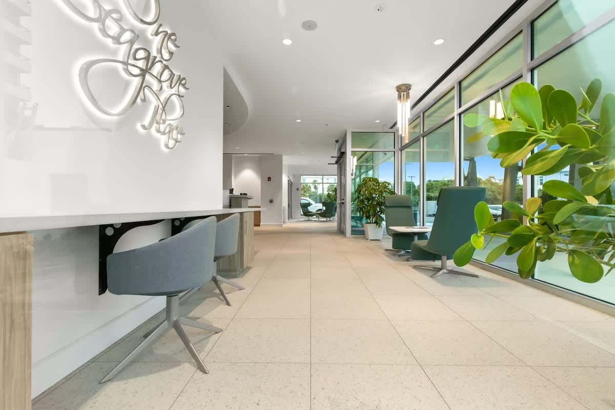 The contemporary lobby offers a long counter with gray swivel stools along one wall, opposite a seating area with stylish green armchairs. A "One Seagrove Place" neon sign glows on the wall.