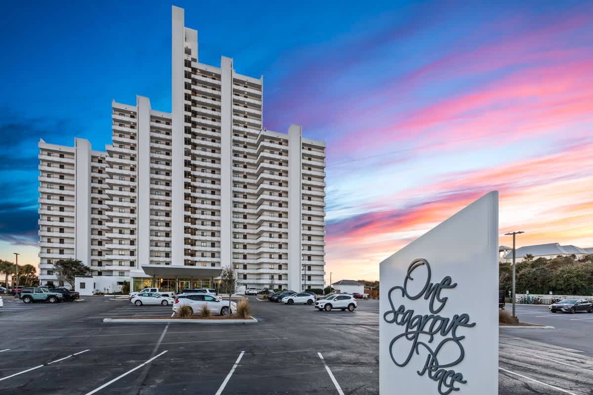 A tall, white condominium building with many balconies stands against a vibrant sunset sky, featuring pink and blue hues. A large parking lot and a prominent "One Seagrove Place" sign are in the foreground.