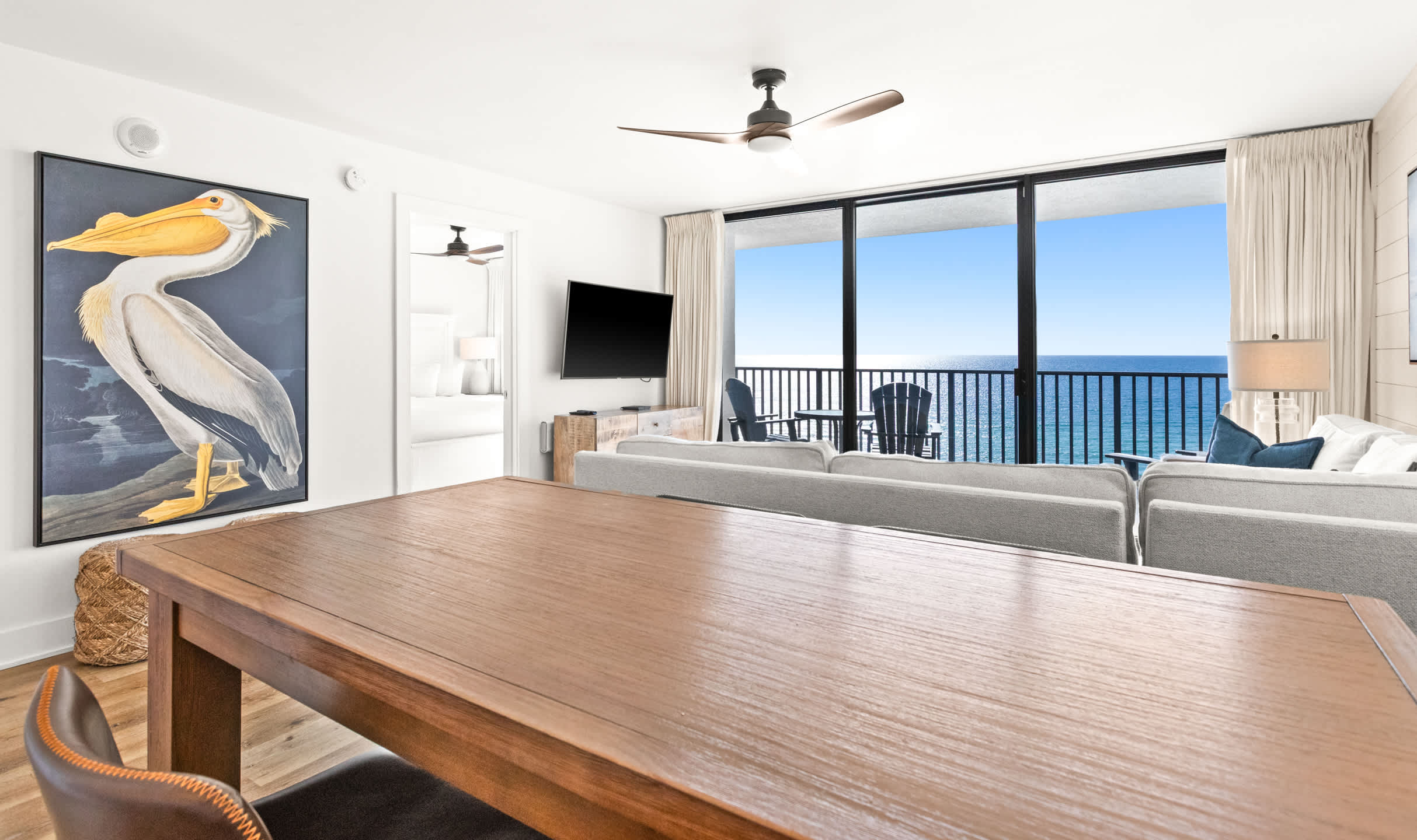 Bright living area with a light gray sectional sofa facing a large flat-screen TV and floor-to-ceiling glass doors opening to an ocean-view balcony, adjacent to a wooden dining table.