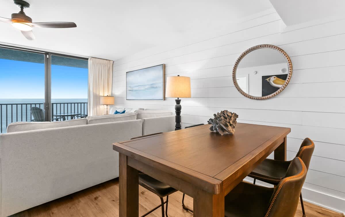 Dining area with a wooden table and brown leather chairs, adorned with a decorative shell, and a round mirror on a shiplap wall, overlooking the living room and ocean.