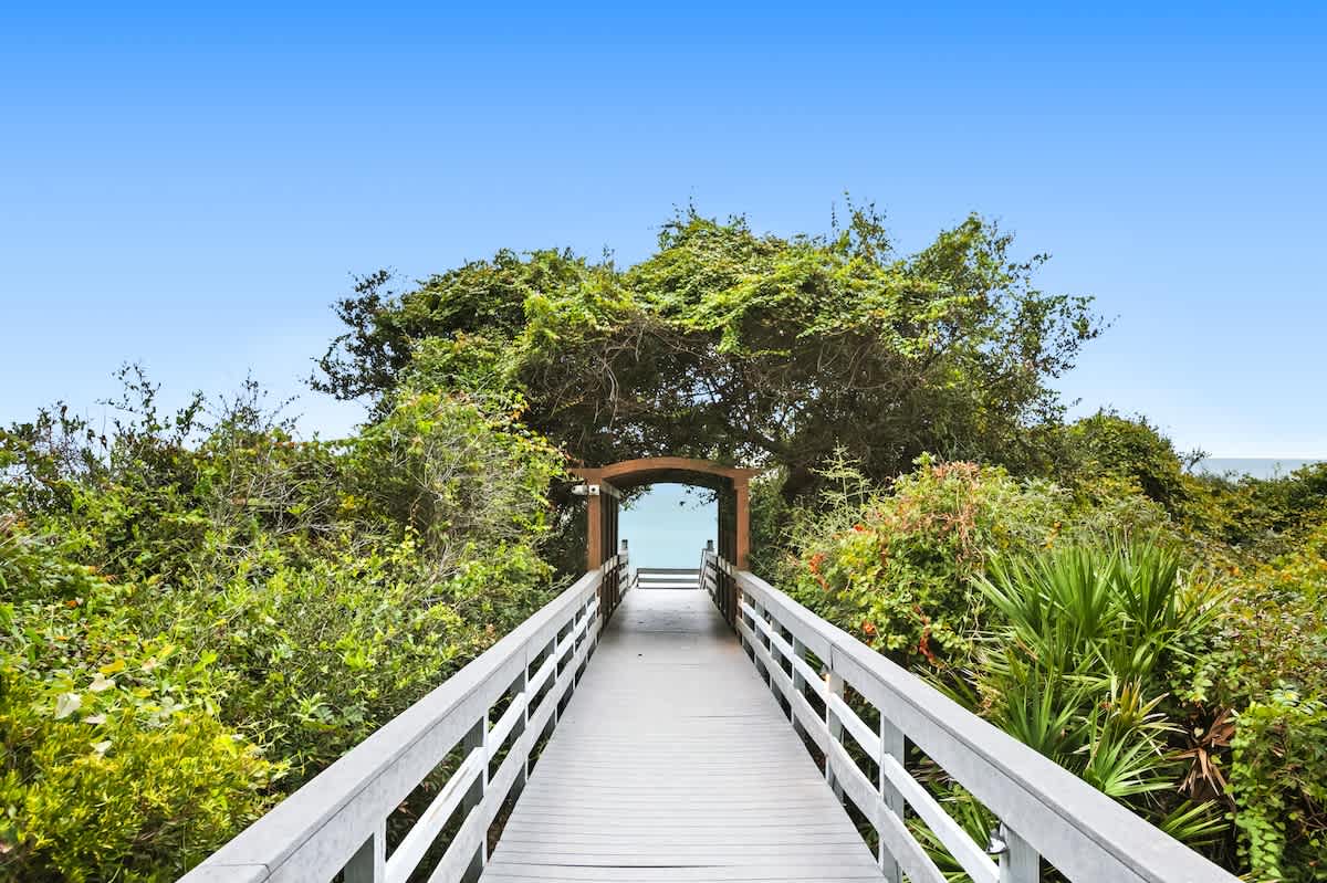 A long wooden boardwalk with railings, winding through lush green coastal foliage, leading towards a glimpse of the blue ocean in the distance.