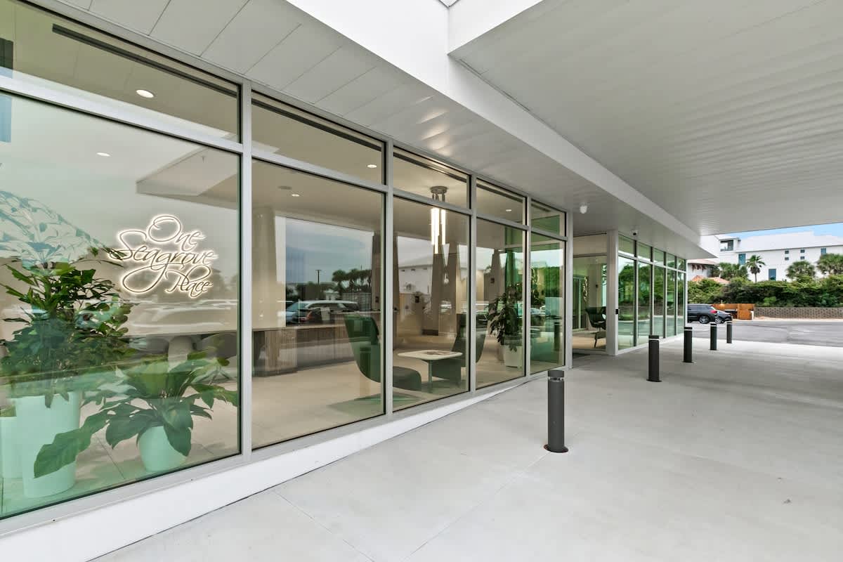 The bright, modern lobby features floor-to-ceiling glass windows offering views outside, complemented by lush indoor plants. A stylish "One Seagrove Place" neon sign adorns a white wall.