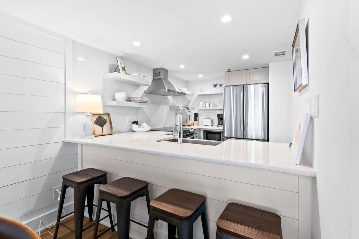 Modern kitchen featuring white subway tile backsplash, stainless steel appliances, open shelving, and a white quartz breakfast bar with four dark wood and metal stools.