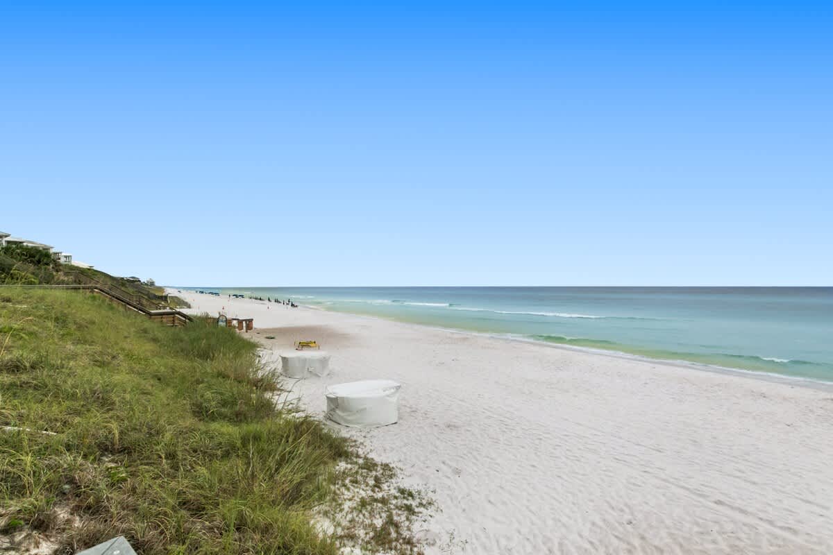 A wide, expansive white sandy beach stretches along the coastline, meeting the clear blue-green ocean under a bright sky. Green grassy dunes line the edge of the beach, with some covered beach equipment visible.