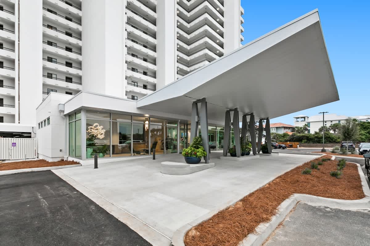 The modern entrance of the condominium building features a striking angular canopy supported by dark pillars, leading to a glass-fronted lobby. Landscaping with pine straw borders the concrete walkway.