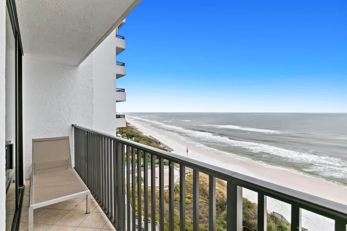 An expansive balcony provides a sweeping view of the coastline, with the ocean stretching to the horizon and a long white sandy beach below. A light-colored lounge chair is visible on the tiled floor, next to a black metal railing.