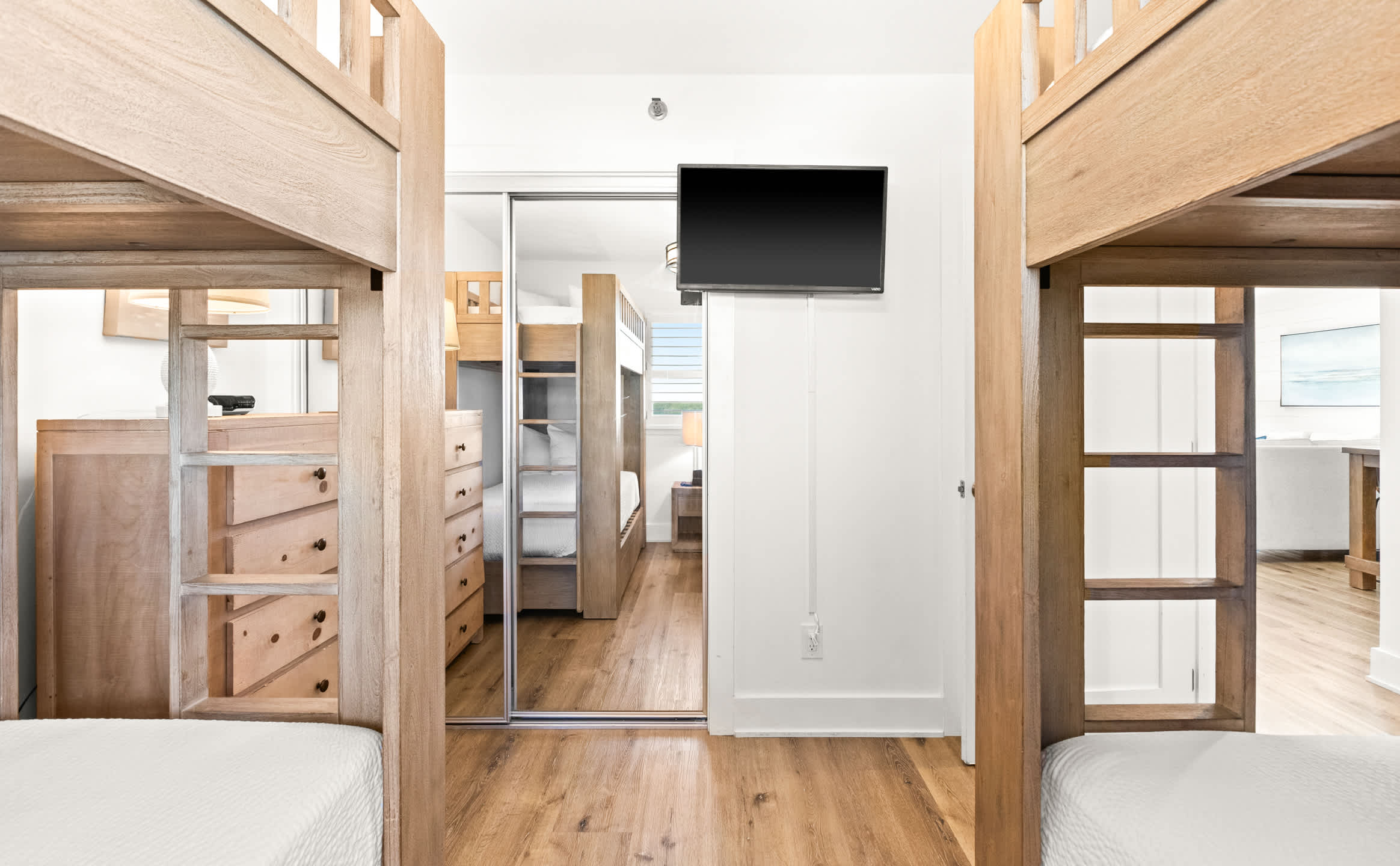 A bright bunk room features two wooden bunk beds with white bedding, separated by a mirrored closet door reflecting another bunk bed. A wall-mounted TV is visible above the closet, and the room has light wood-look flooring.