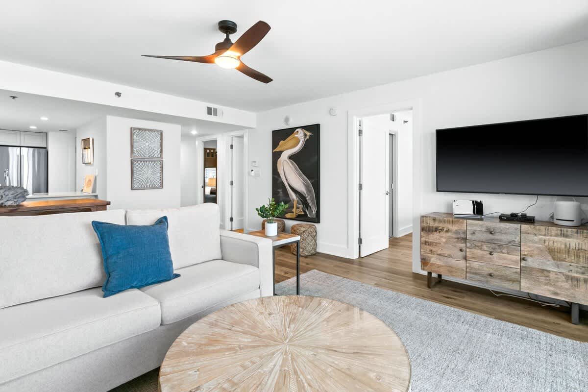 A bright living room showcases a large pelican artwork above a side table, a light-colored sofa, and a rustic wooden console beneath a wall-mounted television, with hardwood floors throughout.