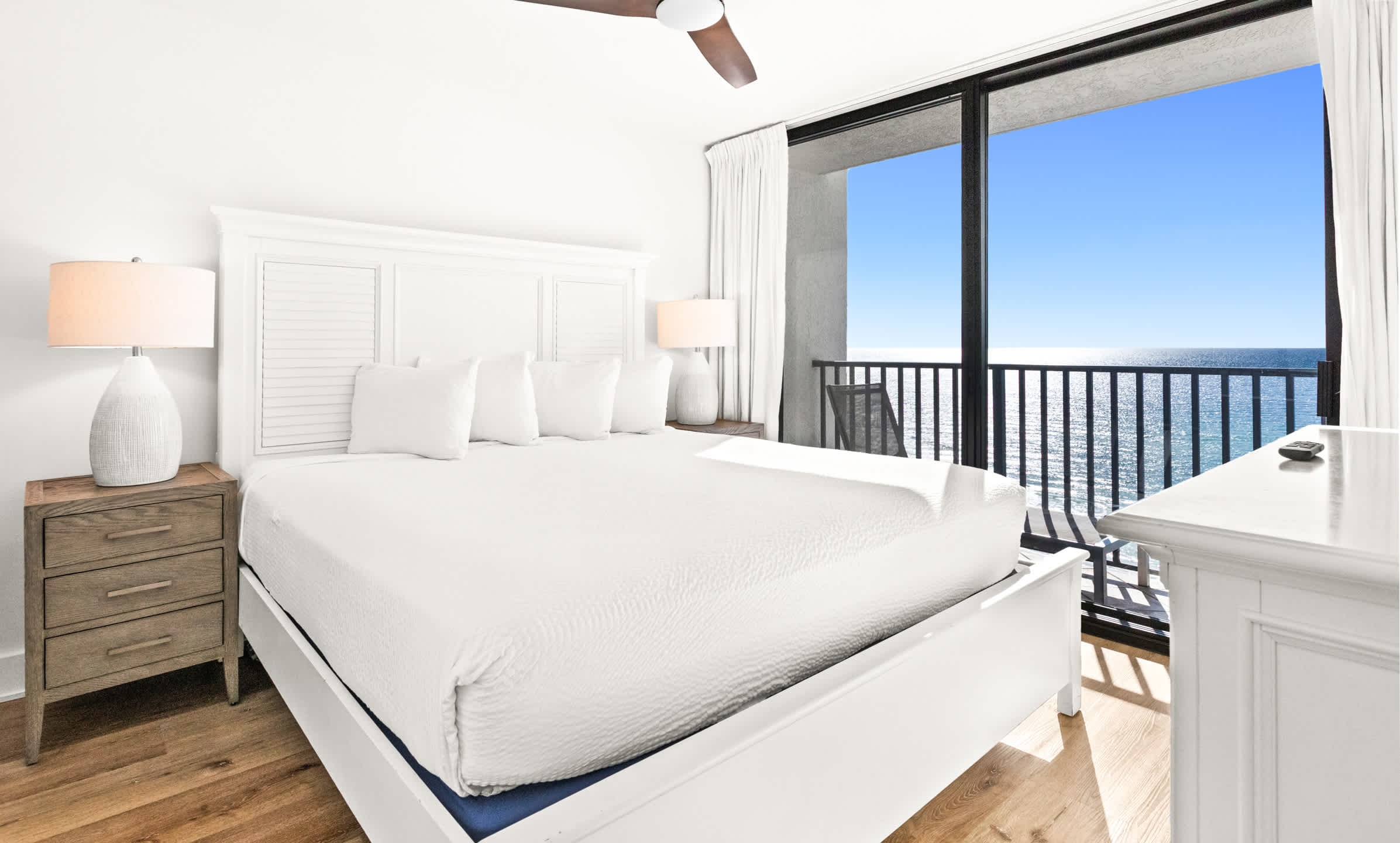 Serene bedroom featuring a white king-sized bed with a slatted headboard, two nightstands with lamps, and a private balcony offering expansive ocean views.