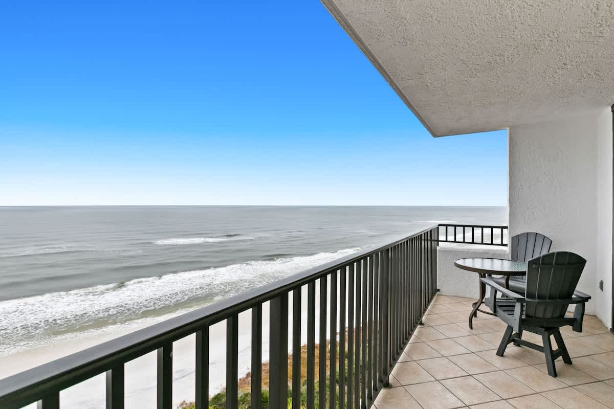 A wide balcony overlooks the vast ocean and a pristine sandy beach, furnished with a glass-top table and two dark Adirondack-style chairs. The balcony features light tiled flooring and a sturdy black metal railing.