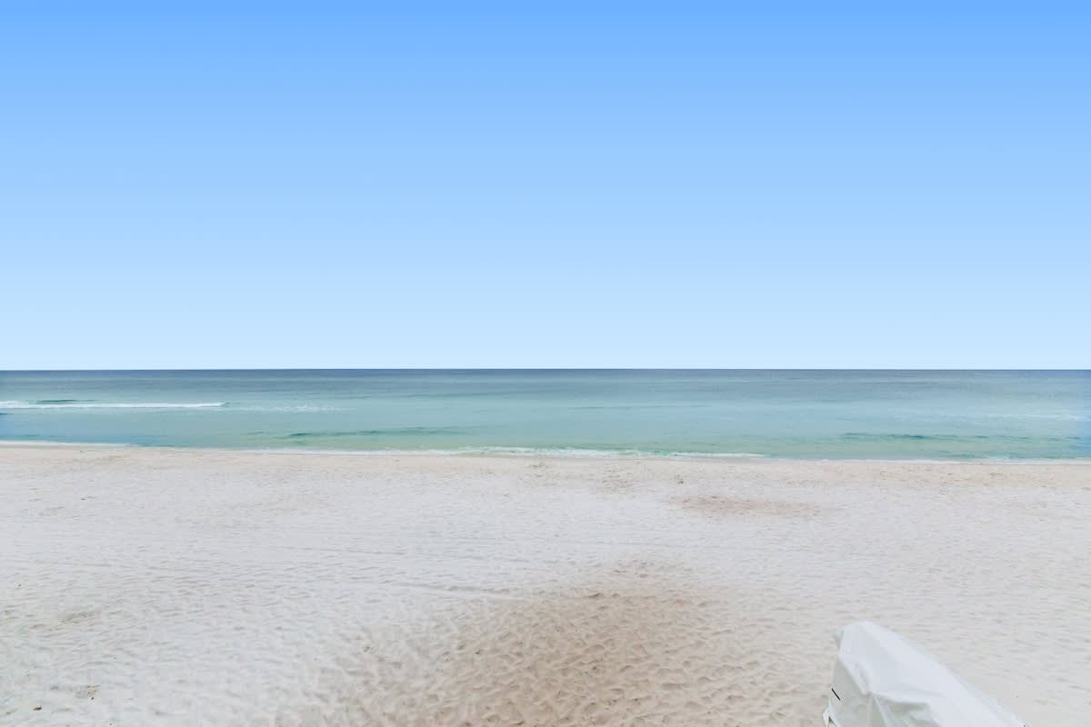 A serene view of a wide, untouched white sandy beach meeting the calm, turquoise ocean under a clear blue sky. The horizon is perfectly flat, emphasizing the vastness of the sea.