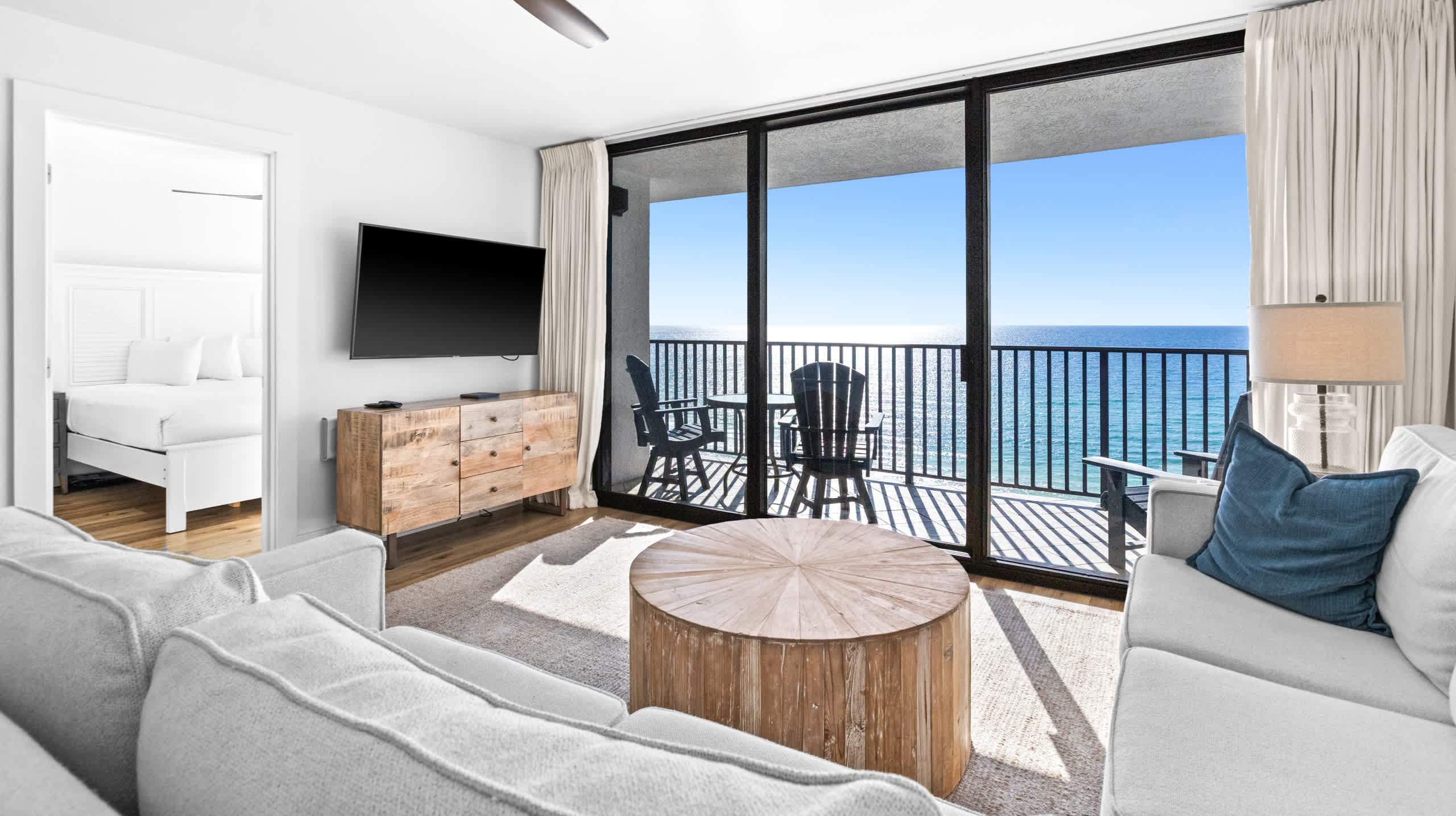 A cozy living room is furnished with a light-toned sectional sofa, a unique round wooden coffee table, and large glass doors providing direct access to a balcony with ocean vistas. A peek into a bedroom is visible to the left.