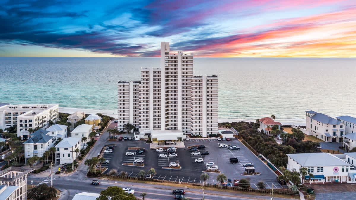 Wide aerial view showcasing a tall white condominium building with multiple balconies, a large parking lot, and surrounding residential properties, all set against the backdrop of the vast blue ocean and a colorful sky.