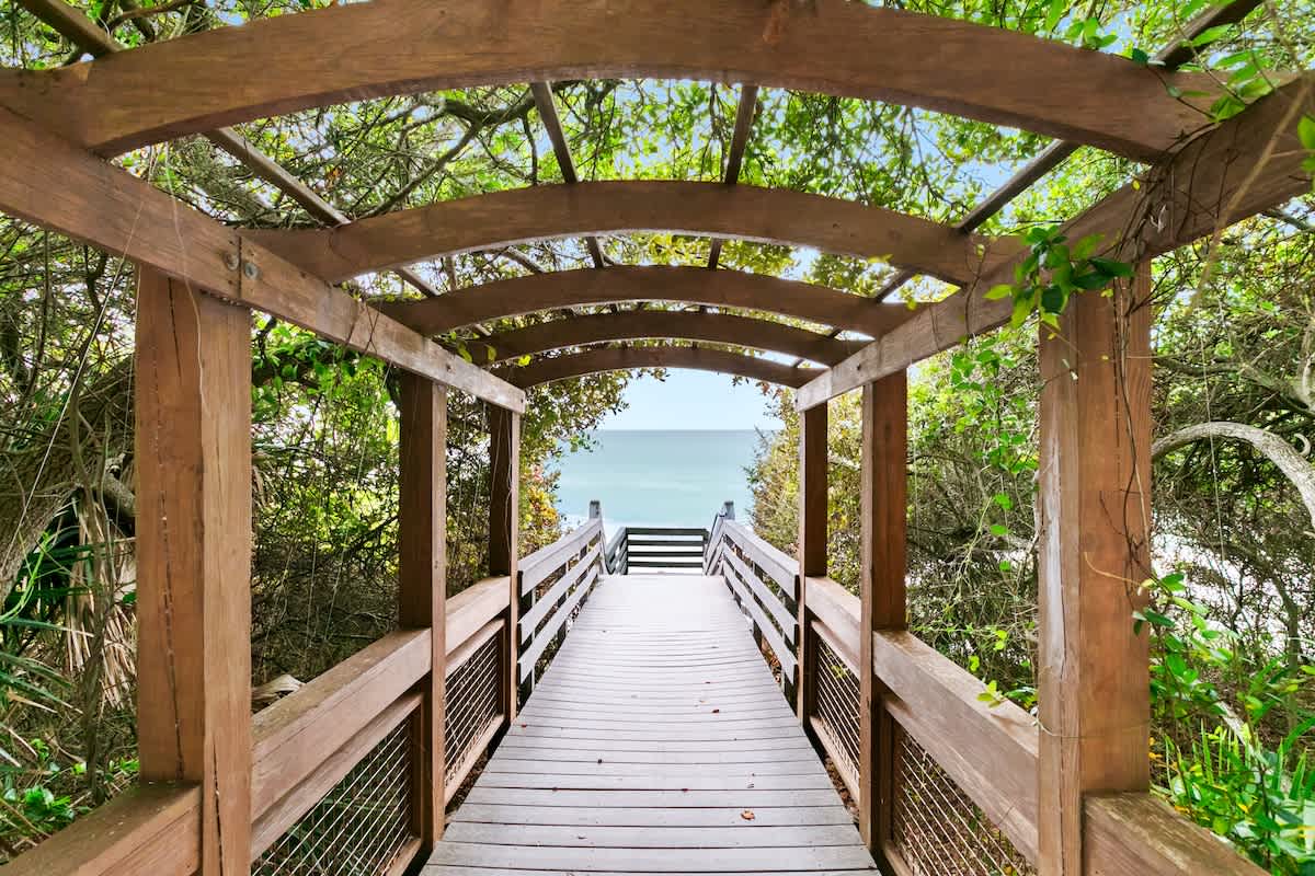 A wooden boardwalk with railings and an arched wooden trellis overhead, surrounded by dense green foliage, leading down towards the visible blue ocean.