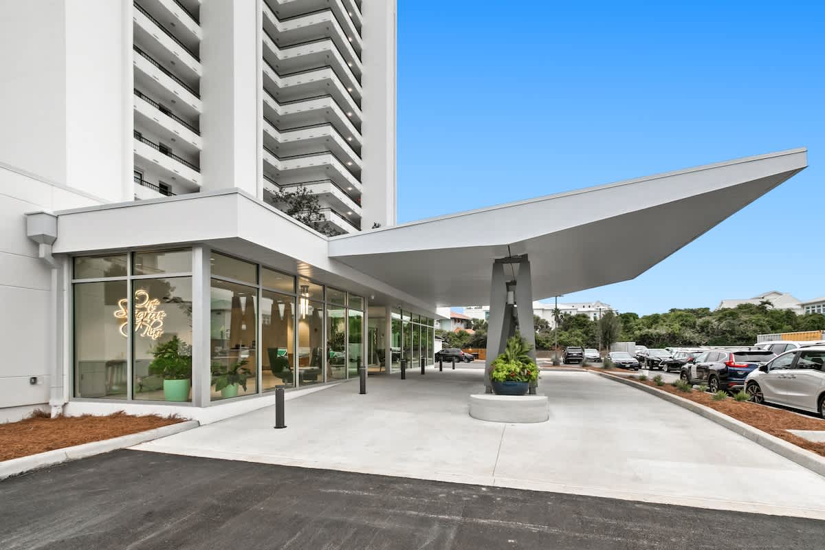 A contemporary building entrance boasts a large, angled white canopy extending over the drop-off area, with expansive glass windows revealing the interior lobby. A paved driveway and parking lot are visible to the right.