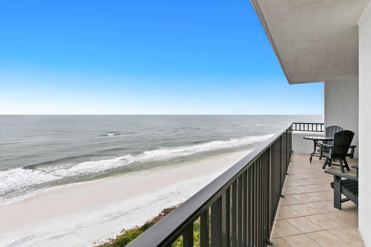 A spacious balcony offers panoramic views of the calm ocean and a wide sandy beach, featuring a small round glass-top table with two dark Adirondack-style chairs. The balcony has light tiled flooring and a black metal railing.