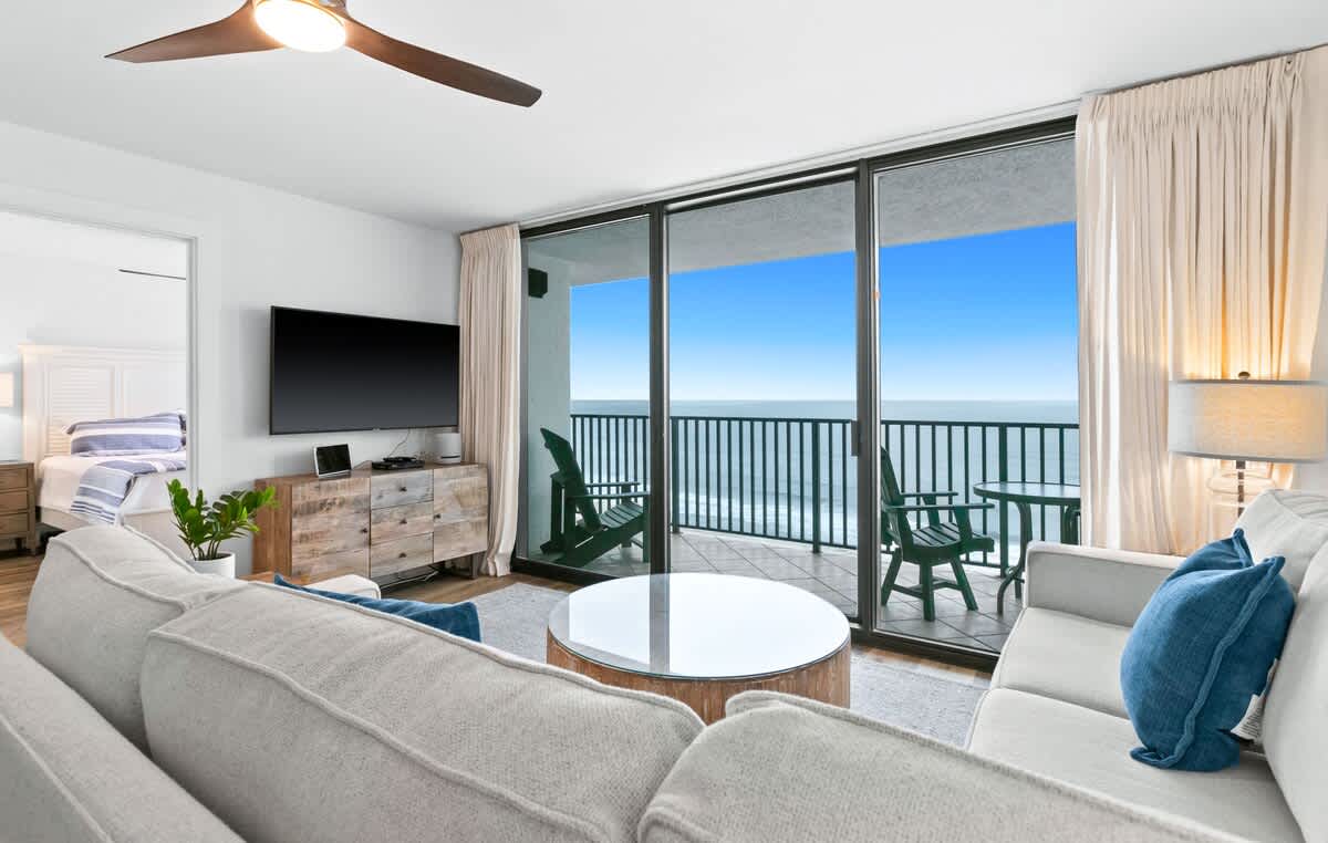 This inviting living room offers a comfortable light-colored sofa, a round wooden coffee table, and expansive sliding glass doors leading to a balcony with chairs and an unobstructed view of the blue ocean.