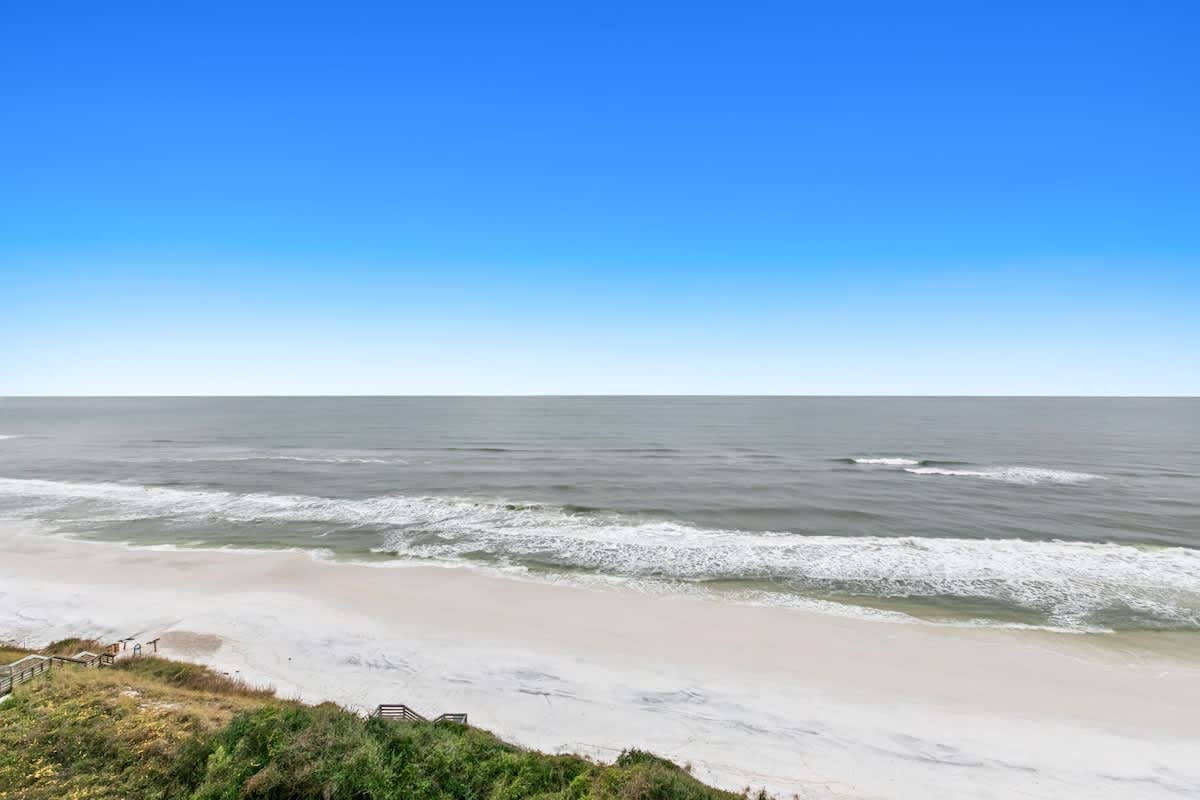 A breathtaking view of the vast, calm ocean with gentle waves breaking on a wide, pristine white sandy beach. Lush green dunes with wooden beach access stairs are visible in the foreground.