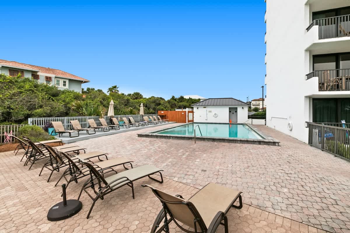 An outdoor swimming pool is surrounded by a large paved patio with numerous lounge chairs, set beside a tall white building and lush green landscaping under a bright blue sky.