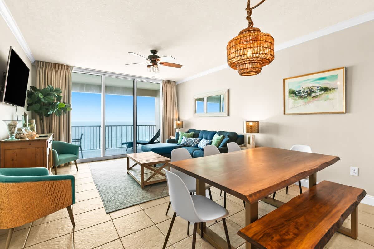 Expansive living and dining area with a live-edge wooden dining table and a blue sectional sofa. Large sliding glass doors open to a balcony, offering panoramic ocean views, complemented by a woven pendant light and ceiling fan.