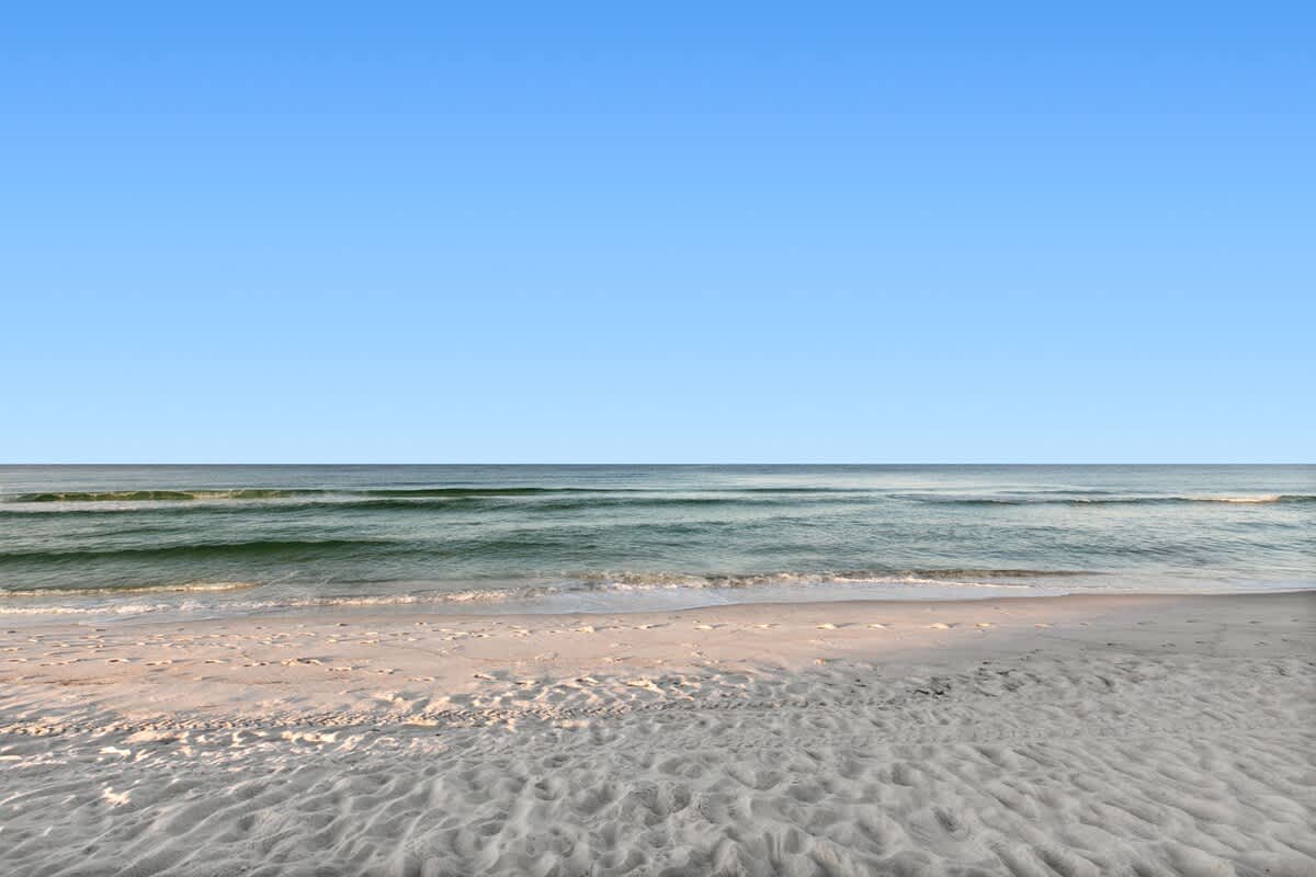 Gentle waves roll onto a pristine white sand beach, creating a serene coastal scene under a bright blue sky. The water transitions from clear to a deeper blue further out.