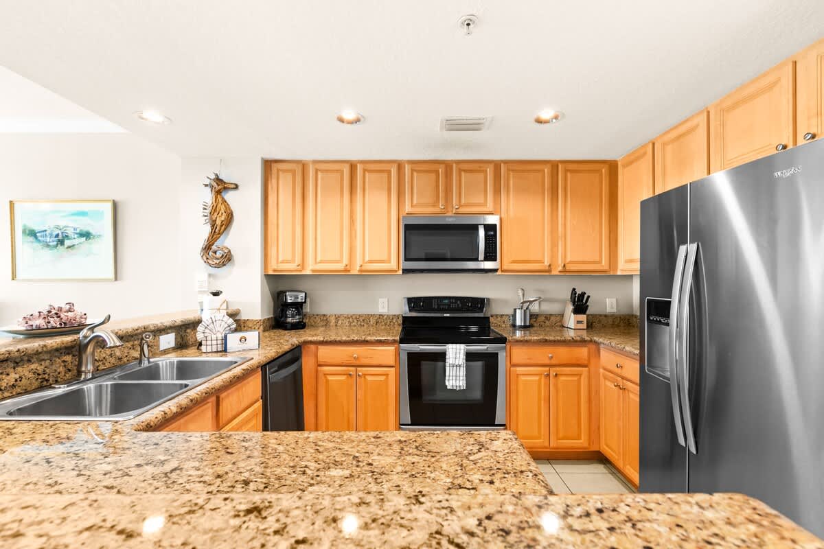 A full view of the kitchen showcasing light wood cabinets, speckled granite countertops, and stainless steel appliances including a refrigerator, microwave, and electric range.