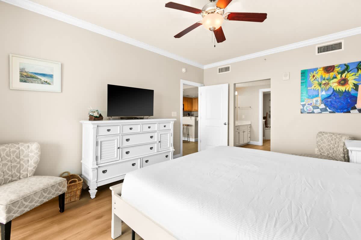 A view of the bedroom from the foot of the bed, showing a white dresser with a TV, an accent chair, and an open doorway leading to an en-suite bathroom.