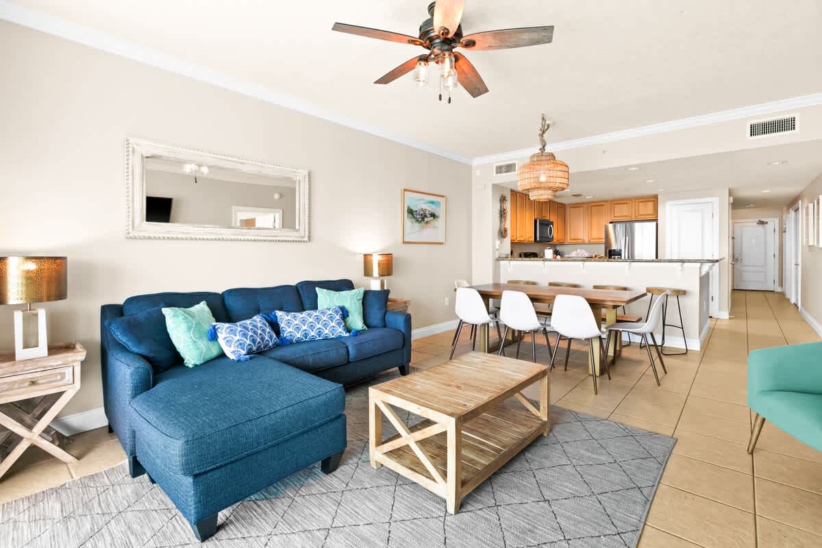 An open-concept living area with a navy blue sectional sofa, a light wood coffee table, and a dining table with white chairs, all set on light tile flooring with a geometric rug.