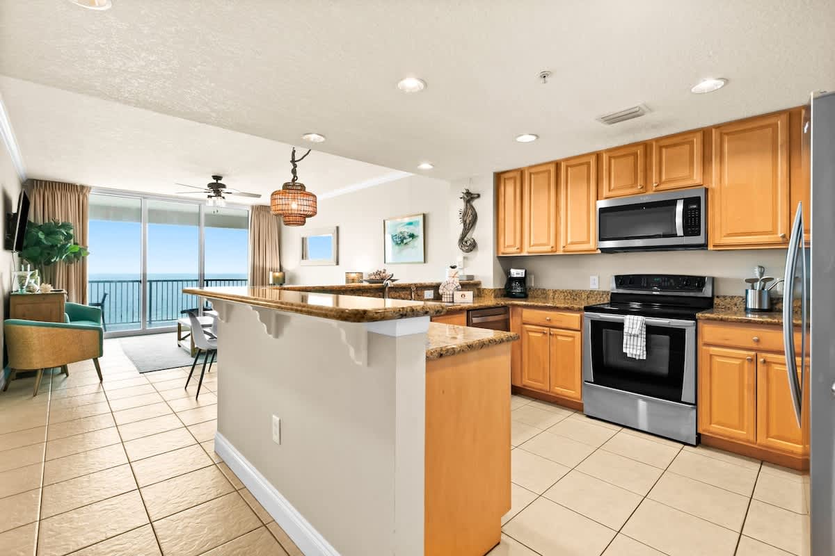Open-concept kitchen with light wood cabinets, granite countertops, and stainless steel appliances, including a dishwasher. It seamlessly connects to the living area, offering glimpses of the ocean through the balcony doors.