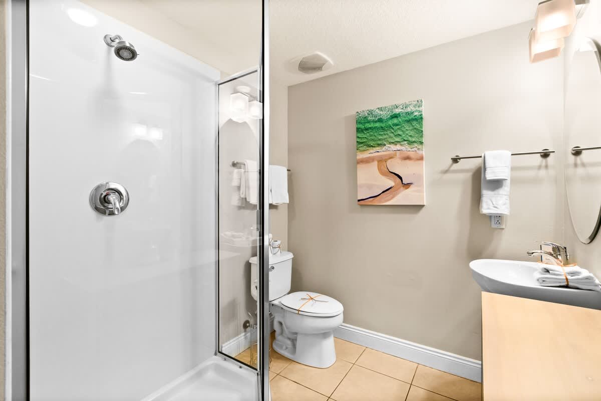 A modern bathroom features a glass-enclosed shower, a white toilet, and a pedestal sink with a round mirror above it. A vibrant beach-themed artwork hangs on the wall.