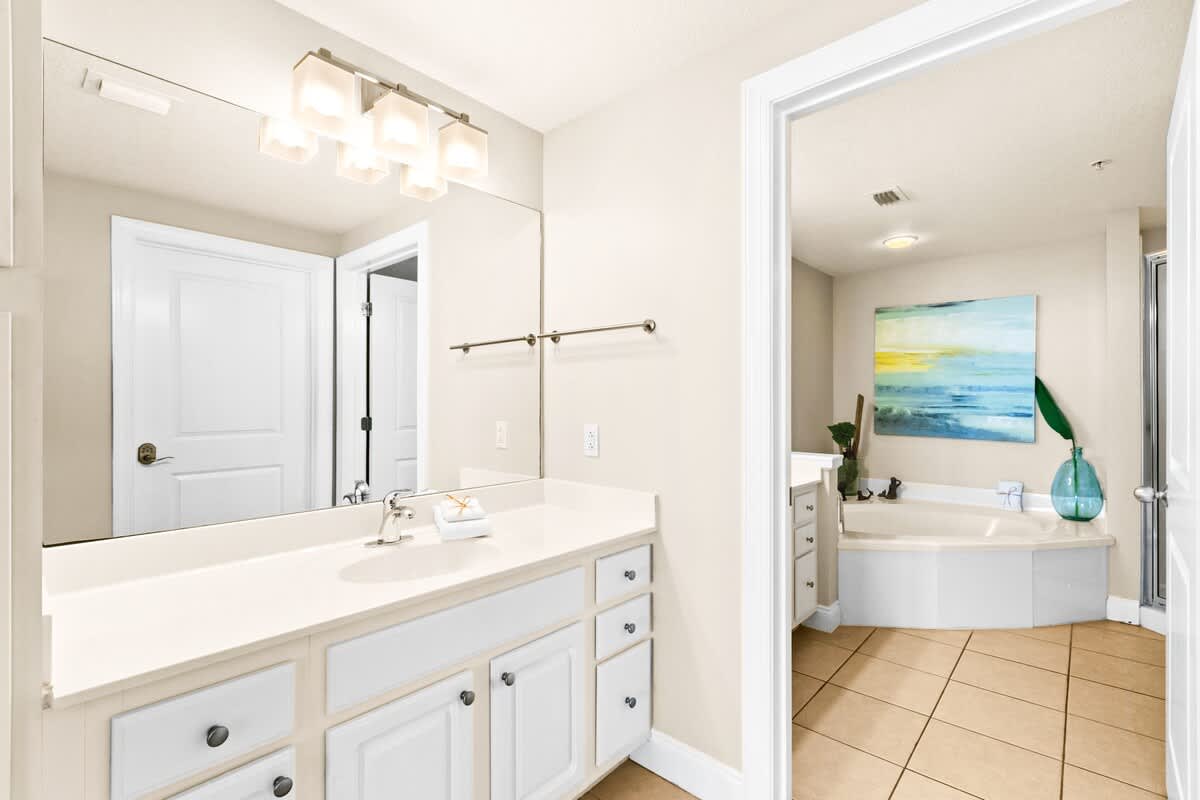 A modern bathroom vanity with a single sink, white cabinets, and a large mirror, with a glimpse of a jetted tub and shower in the background.
