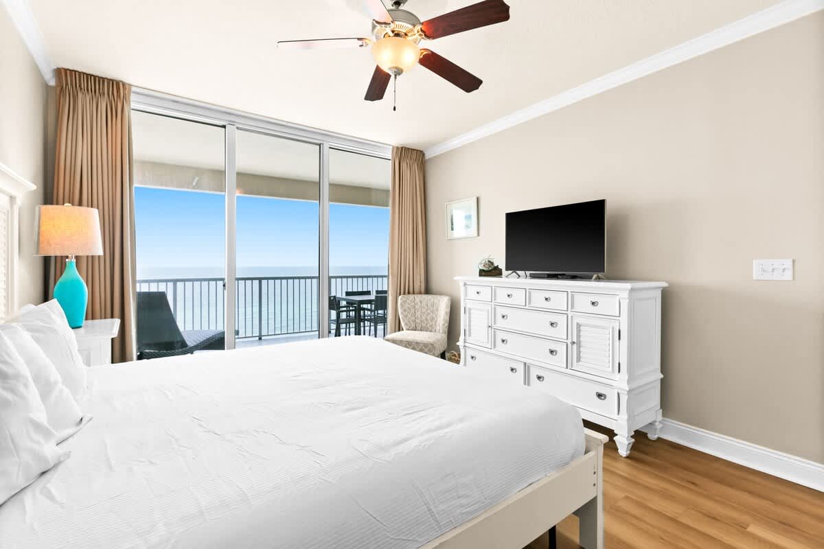 A spacious bedroom with a white bed frame, matching dresser, and a flat-screen TV, featuring light wood flooring and large sliding glass doors leading to a balcony with ocean views.