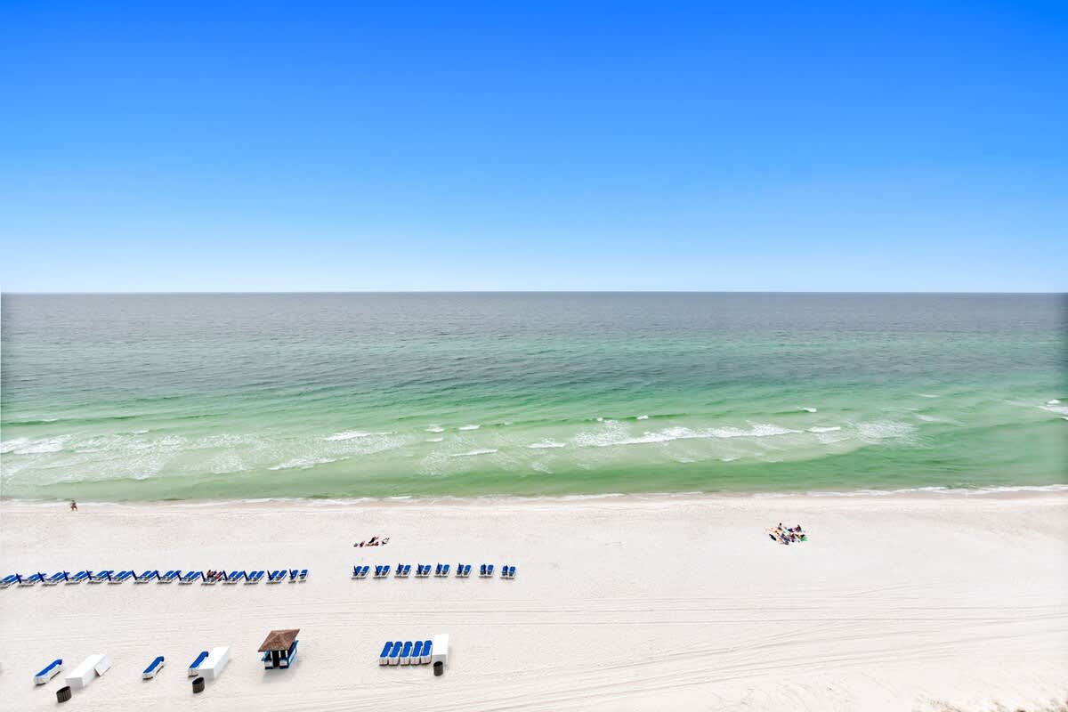 A stunning aerial view captures the expansive white sand beach and the clear turquoise waters of the ocean. Numerous beach chairs and umbrellas are set up on the sand.