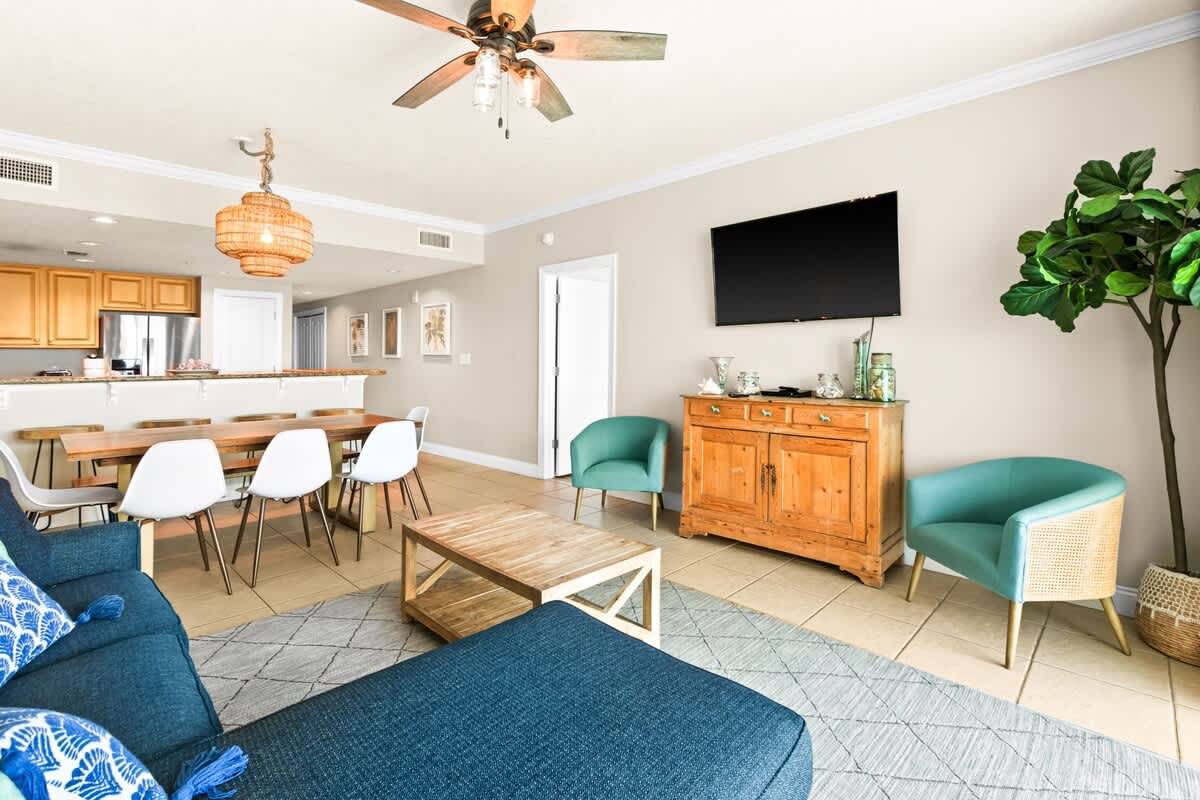 Open-plan dining and living area showcasing a live-edge wooden dining table with white chairs and a bench. A blue sectional sofa and wooden coffee table define the living space, with the kitchen bar visible in the background.