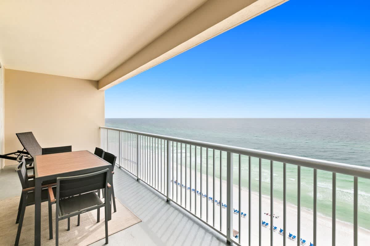 A spacious balcony offers panoramic views of the turquoise ocean and white sandy beach below. It is furnished with an outdoor dining table and chairs.